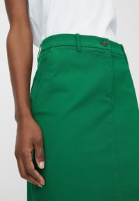 Gros plan sur une personne portant un pantalon vert avec des poches avant et un bouton noir, associé à un haut blanc.