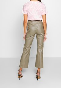 Kvinna i högmidjade taupe läderbyxor, rosa tie-dye T-shirt och högklackade sandaler med ormskinnsmönster, som står mot en enfärgad bakgrund.