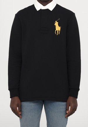 Man draagt een zwart poloshirt met lange mouwen, een witte kraag en een goudkleurig geborduurd polospelerlogo op de borst, gecombineerd met een blauwe spijkerbroek.