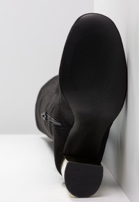 Botte d'archive en daim noir avec fermeture éclair latérale, montrant une semelle en caoutchouc texturée et un talon épais de hauteur moyenne sur fond blanc.