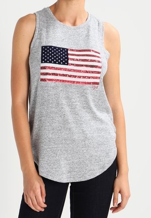 Szary top bez rękawów z cekinową flagą USA na piersi, noszony przez osobę w ciemnych spodniach, stojącą na białym tle.