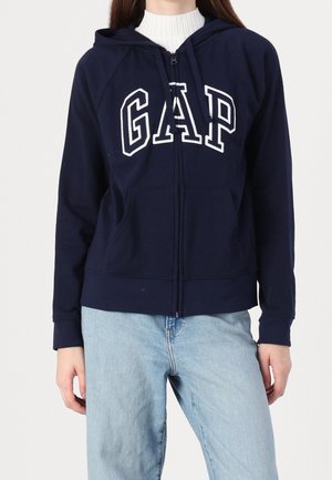 Femme portant un sweat à capuche zippé bleu marine avec le lettrage "GAP" et un jean bleu clair, visible du cou aux hanches sur un fond blanc.