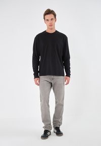 Långärmad svart skjorta, grå jeans och svarta skor med vita ränder. Enkelt mönster, slät textur, står framför en enfärgad bakgrund.