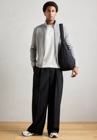 Γκρι ζακέτα με φερμουάρ πάνω από ένα λευκό πουκάμισο, συνδυασμένη με χαλαρές μαύρες παντελόνες και λευκά sneakers. Το μοντέλο κρατά μια μαύρη τσάντα χιαστί. Απλό φόντο.