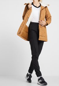 Brązowa parka z białym futerkiem w środku, z zapinanymi na suwak kieszeniami i zapięciami na zatrzaski, zestawiona z czarnymi spodniami sportowymi i czarnymi trampkami.