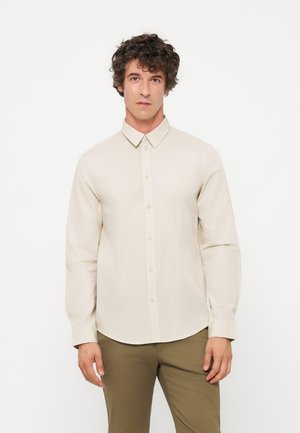 Homme aux cheveux bouclés portant une chemise beige à manches longues boutonnée et un pantalon olive, debout devant un fond blanc uni.