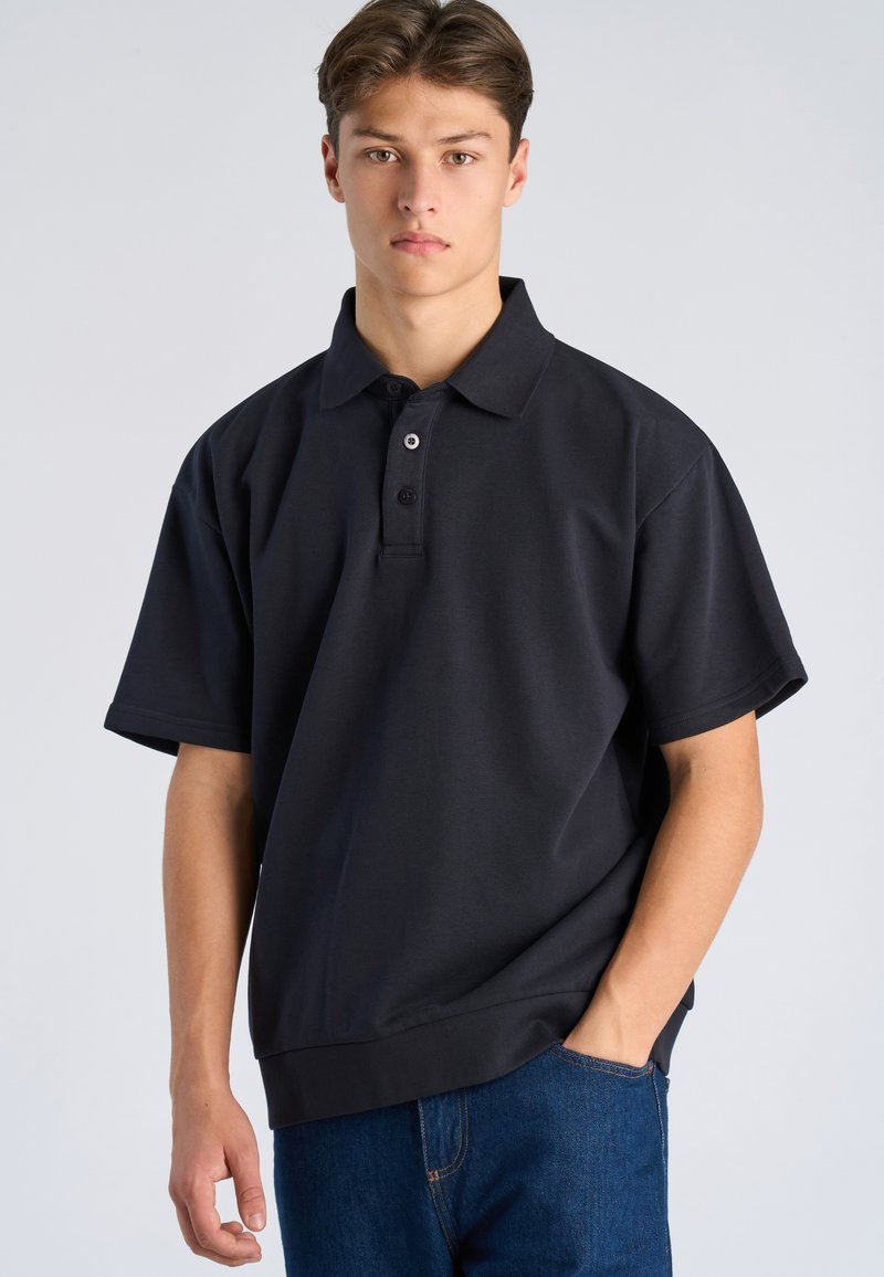 Jonge man met bruin haar, gekleed in een donkerblauw poloshirt met korte mouwen en een blauwe jeans, staand met één hand in zijn broekzak tegen een effen achtergrond.