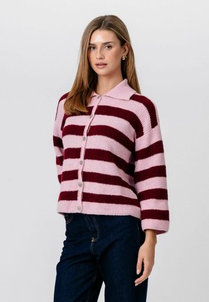 Femme aux cheveux lisses portant un pull à rayures roses et bordeaux à boutons et un jean bleu foncé, debout devant un fond clair.