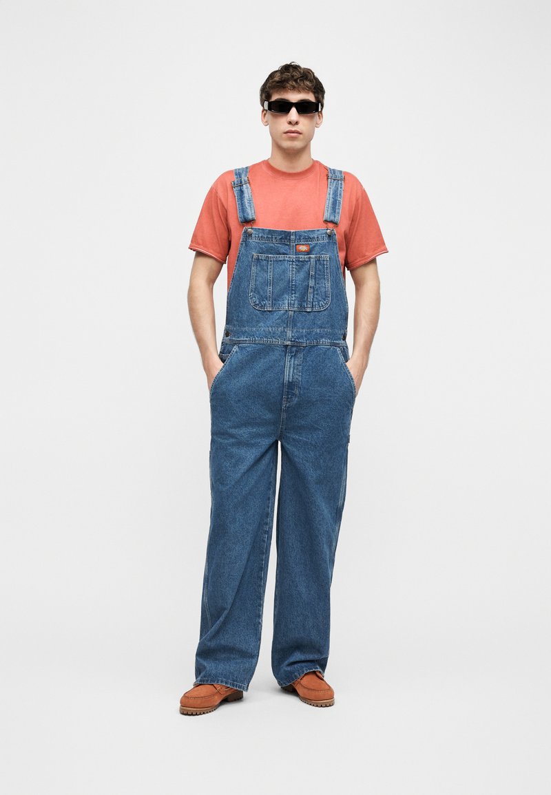 Giovane uomo con occhiali da sole neri, una maglietta rossa sbiadita, salopette di denim blu e scarpe marroni, in piedi con le mani in tasca.
