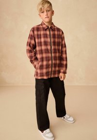 Rød og sort ternet lynlåsjakke med to sidelommer, parret med sorte cargo-bukser og hvide sneakers med grå og brune detaljer.