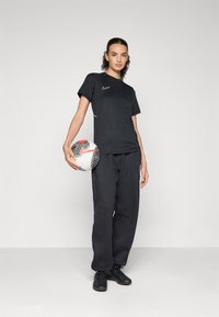 Svart Nike T-skjorte med subtil logo og hvite detaljer, kombinert med løse svarte joggebukser. Modellen holder en flerfarget fotball.
