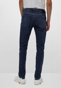 Muži oblečení v tmavě modrých slim-fit džínách, bílých teniskách a bílém tričku, stojící proti jednobarevnému světlému pozadí.