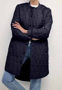 Cappotto lungo trapuntato blu navy con motivo a diamante, bottoni a pressione e polsini a coste. Indossato sopra una camicia bianca e jeans blu.