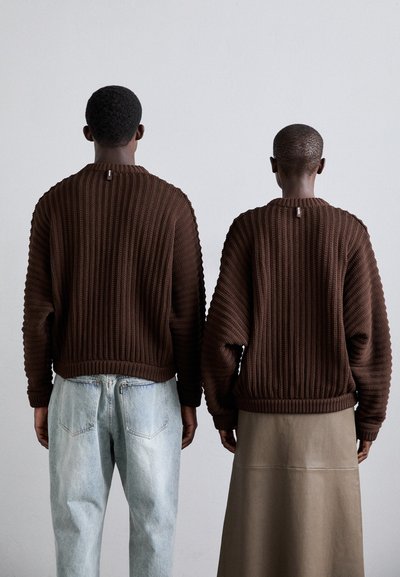 Suéteres de punto texturizados en marrón con diseño de canalé, llevados sobre jeans de un azul claro y una larga falda beige. Ambos se ven desde atrás.