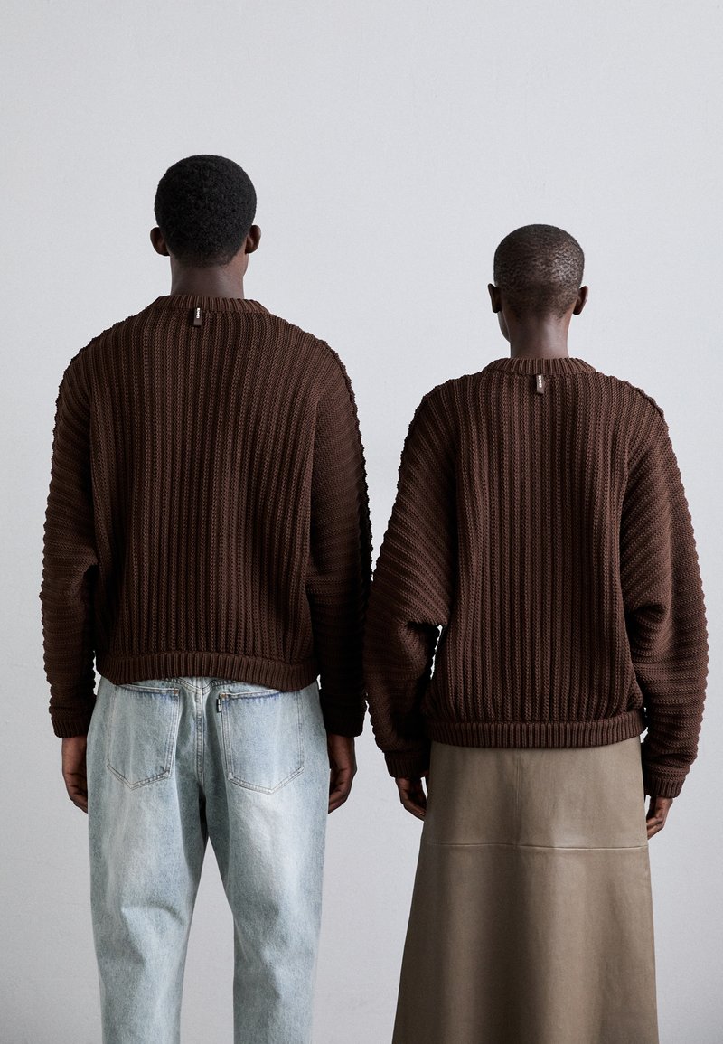 Brown textured knit sweaters with ribbed design, worn over light blue jeans and a long beige skirt. Both are viewed from the back.
