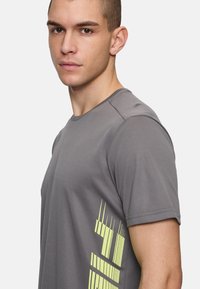 Camiseta atlética gris hecha de material transpirable con mangas cortas. Presenta un diseño geométrico en neón amarillo en el lado izquierdo.