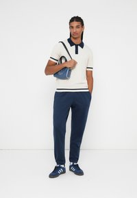 Camiseta polo crema con ribetes en azul marino, combinada con pantalones de chándal azul marino y zapatillas azules. Los accesorios incluyen un pequeño bolso azul con detalles de logo.