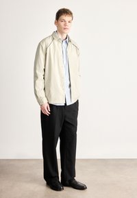 Casaco de camisa bege claro com costuras pretas, usado sobre uma camisa de botão branca com riscas azuis, combinado com calças largas pretas e sapatos de couro.