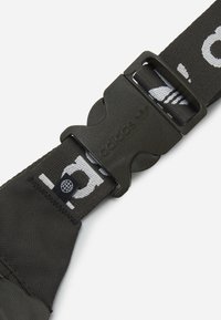 Černý nastavitelný popruh s plastovou sponou, na které je vyražené logo "adidas". Tkanina má bílé vzory na texturovaném pozadí.