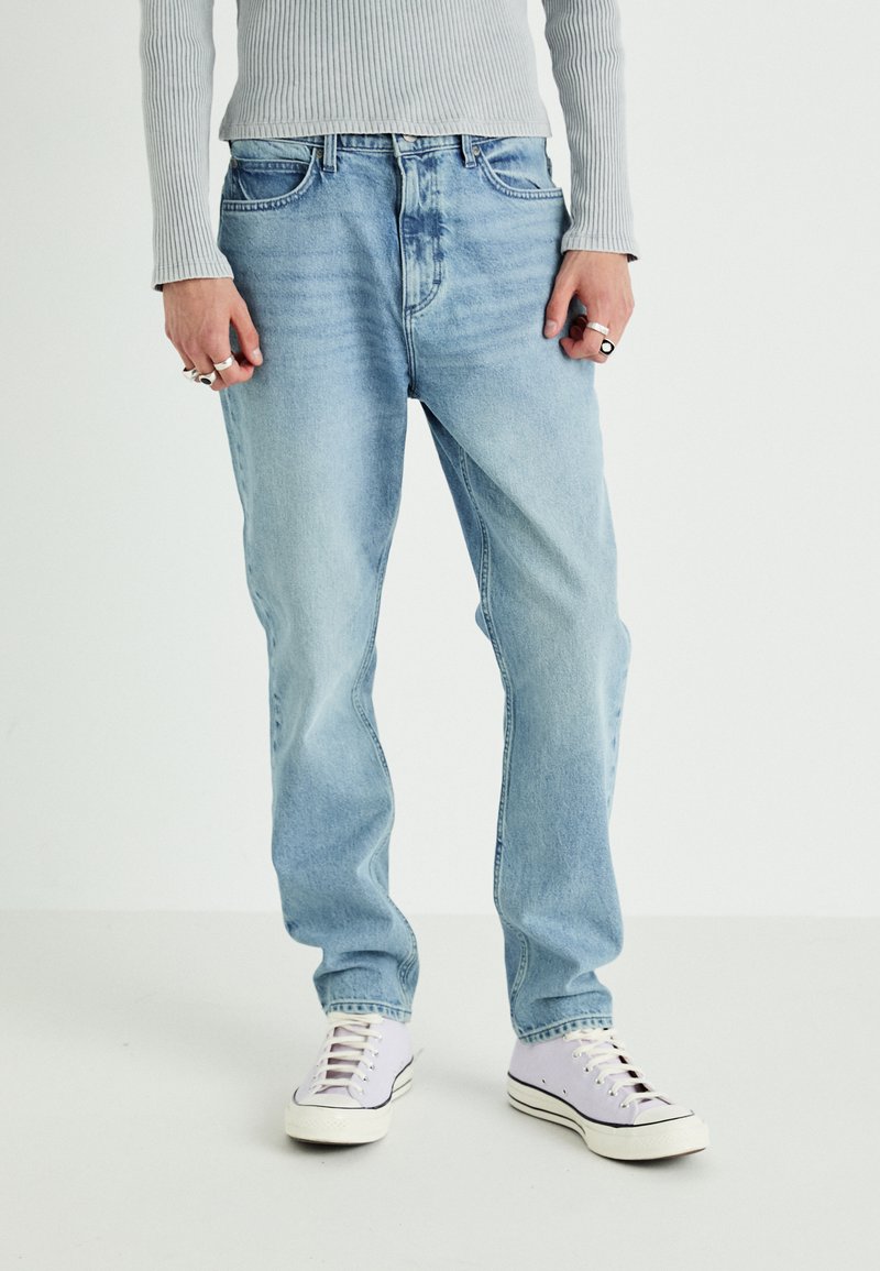 Svetlo modri jeans s ravnim krojem, petimi žepi, v kombinaciji s sivo rebrastim dolgim topom in belimi športnimi copati.