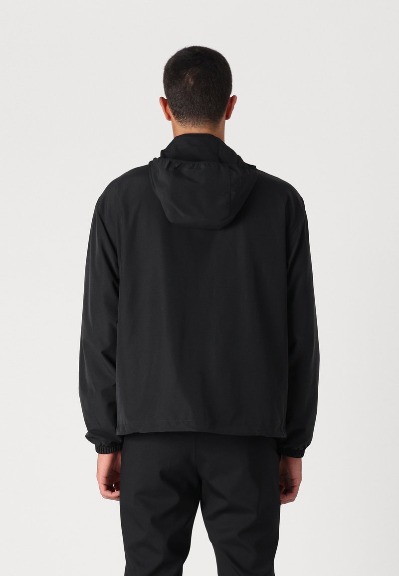 Veste légère noire avec capuche, poignets élastiques et coupe décontractée, présentée de dos sur fond blanc uni.