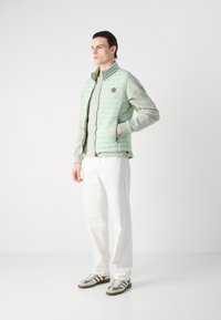 Chaleco acolchado verde claro con patrón de acolchado vertical, cierre con cremallera en la parte delantera y parche del logo, superpuesto a una sudadera a juego y pantalones blancos.