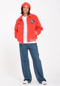 Veste varsity en molleton rouge avec des patchs brodés, des poignets et un col côtelés, assortie à un jean bleu ample et un bonnet rouge assorti.