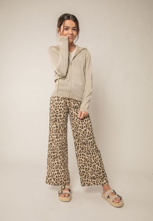 Jong meisje met lang haar, gekleed in een beige hoodie met ritssluiting, wijde broek met luipaardprint en beige sandalen, staand met hand bij het gezicht.