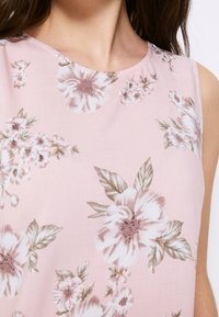 Mouwloze blouse van zachte roze stof met witte en bruine bloemenpatronen. Heeft een ronde hals en een gladde textuur.