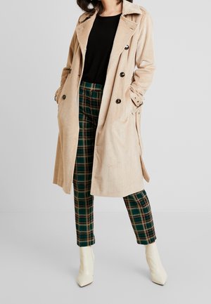 Kobieta w beżowym płaszczu sztruksowym, czarnym topie, zielonych spodniach w kratę i białych botkach, stoi z rękami w kieszeniach płaszcza.