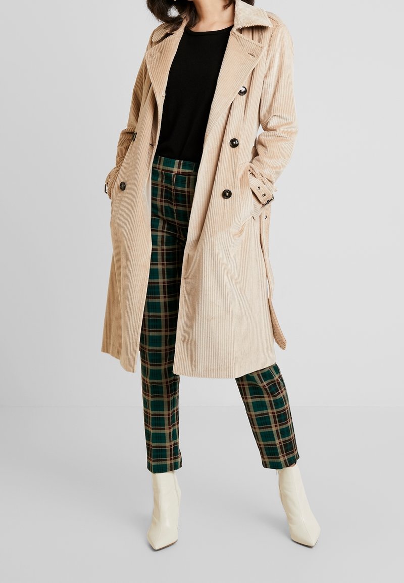 Femme portant un trench-coat en velours côtelé beige, un haut noir, un pantalon à carreaux verts et des bottines blanches, debout les mains dans les poches du manteau.