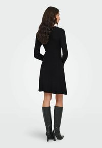 Frau mit langen, welligen Haaren, die ein schwarzes, langärmliges, knielanges Kleid und schwarze, kniehohe Stiefel mit Absatz trägt, seitlich stehend mit den Händen hinter dem Rücken.