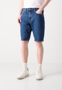 Mørkeblå denimshorts med klassisk snitt, som har lommer og en frayed kant. Kombinert med hvite sneakers og ankellange sokker.