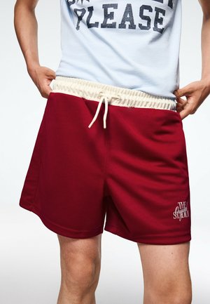 Person trägt rote Shorts mit weißem Kordelbund und weißem Textlogo am linken Bein und justiert den Bund auf beiden Seiten.