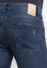 Jeans de mezclilla azul oscuro con corte recto, que cuentan con una etiqueta de cuero en la parte trasera de la cintura y un único bolsillo trasero con un desgaste mínimo.