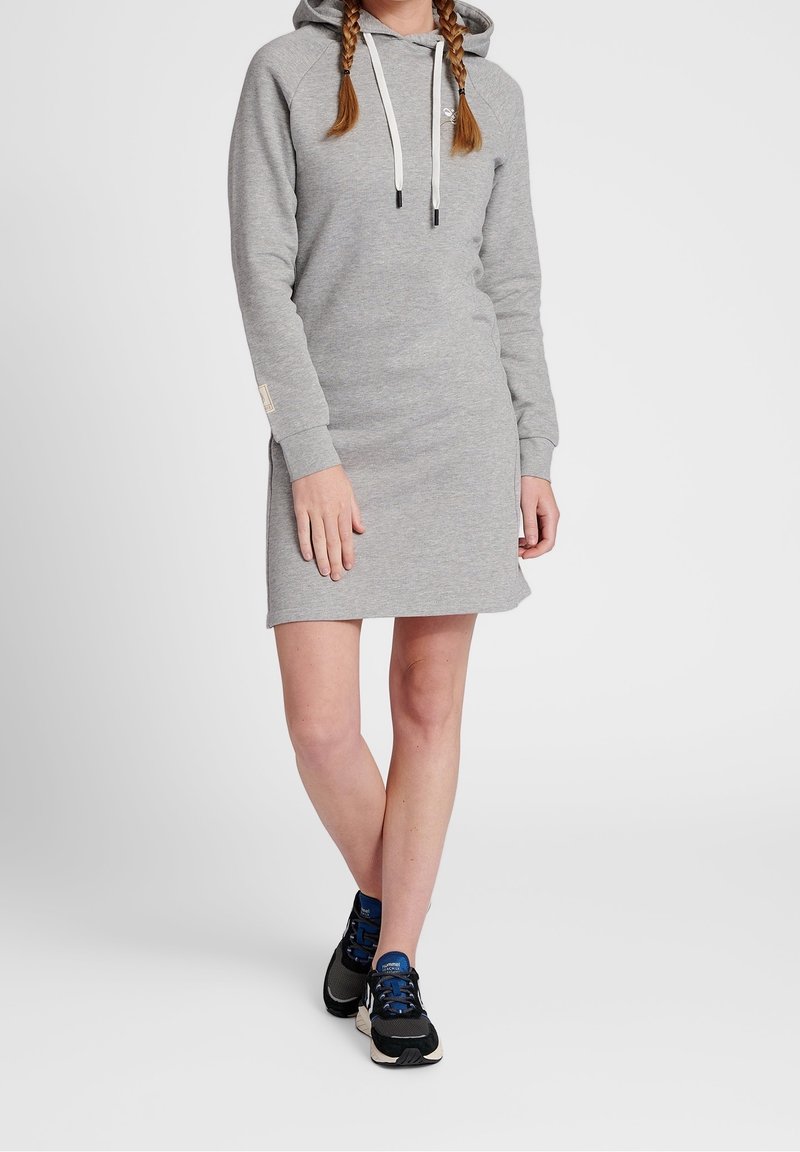 Robe-sweat gris avec capuche, manches longues et fentes sur les côtés. Dotée d'un cordon de serrage avec des embouts noirs. Associée à des chaussures de sport noires.