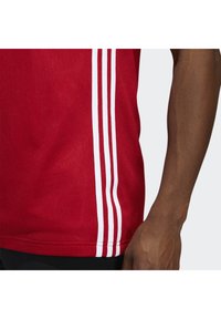 Maillot de sport rouge avec un tissu texturé, des manches courtes et trois rayures blanches sur les côtés. Détail de l'ourlet visible.
