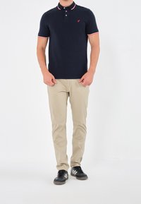 Polo bleu marine avec col rayé rouge et blanc, manches courtes et petit logo rouge. Un pantalon beige et des baskets noires complètent la tenue.