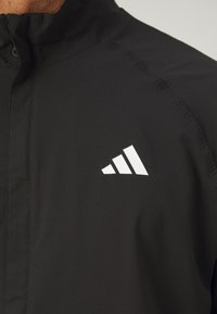 Zwarte sportjas met een hoge kraag, voorzien van een wit driehoekig Adidas-logo op de linkerborst. Glad textuur en lichtgewicht materiaal.