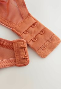 Koralorandžs krūštura siksniņa ar teksturētu audumu, āķiem un cilpām regulēšanai. Mīksta sietveida materiāla detaļas ir redzamas pie piestiprināšanas punktiem.