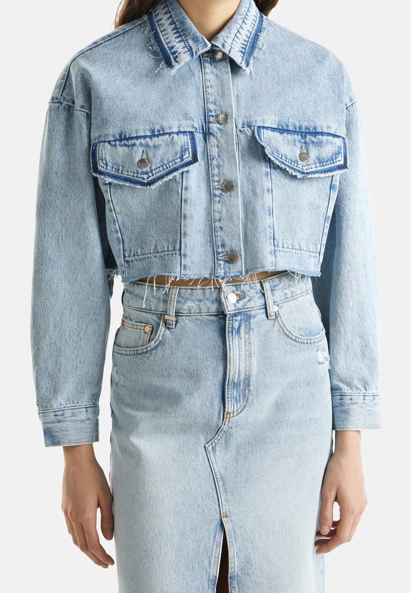 Avkortad ljusblå denimjacka med två bröstfickor, silverknappar och fransade kanter, i kombination med en matchande högmidjad denimkjol.