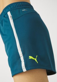 Pantaloni sportivi turchesi con una tasca con zip bianca e un logo Puma giallo. Realizzati in tessuto traspirante che assorbe l'umidità, con una cintura in rete traspirante.