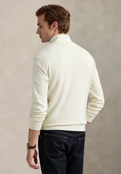 Bărbat cu păr castaniu care poartă un pulover alb cu guler înalt și blugi închiși la culoare, stând cu capul ușor întors spre stânga.