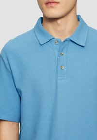 Svetle modrá polo košile z texturované látky, s límečkem, dvěma knoflíky a krátkými rukávy. Žádné viditelné vzory ani loga.
