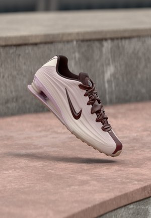 Svetlo bež in rjav Nike copat z vidnim zračno podplatom, ki lebdi nad kamnito površino v zunanjem urbanem okolju.