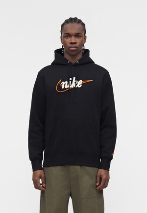 Mann mit geflochtenem Haar, der einen schwarzen Nike-Hoodie mit orange-weißem Logo und olivgrünen Hosen trägt, steht vor einem einfarbigen Hintergrund.