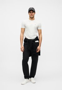 Άνδρας που φοράει λευκό μπλουζάκι, μαύρο παντελόνι, λευκά αθλητικά παπούτσια, μαύρο φούτερ δεμένο γύρω από τη μέση και σκούφο του μπέιζμπολ, στέκεται μπροστά σε απλό φόντο.