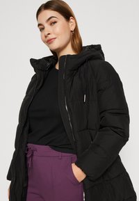 Juodas pūkinis džemperis su gobtuvu ir užtrauktuku priekyje, su kojiniu dizainu. Modelis dėvi juodą raukinį marškinėlį ir purpurinius kelnes.