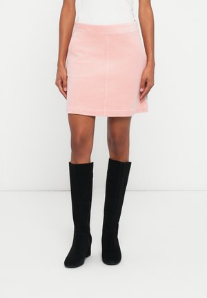 Persoon die een lichtroze corduroy minirok draagt, een wit topje en zwarte kniehoge laarzen, staand tegen een effen witte achtergrond.