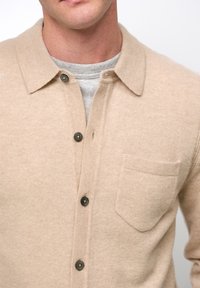 Beige Strickjacke mit Knopfverschluss, dunklen Knöpfen und einer Brusttasche. Weicher Stoff, klassisches Kragen-design, über einem grauen Hemd getragen.